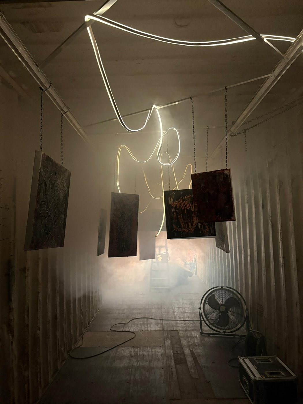 The container in the dark: canvases hanging on chains under a looping neon line, haze across the floor