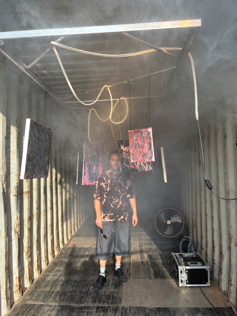 Wessel standing in the container between hanging canvases, daylight and haze at both ends